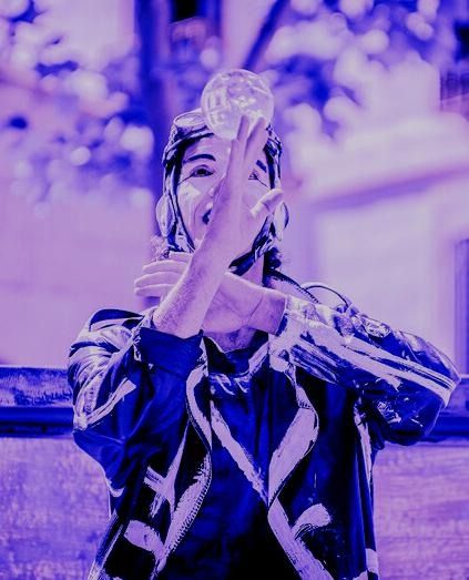 Una persona con ropa estampada azul balancea una pelota transparente sobre la cabeza, con los brazos cruzados. Fondo morado.